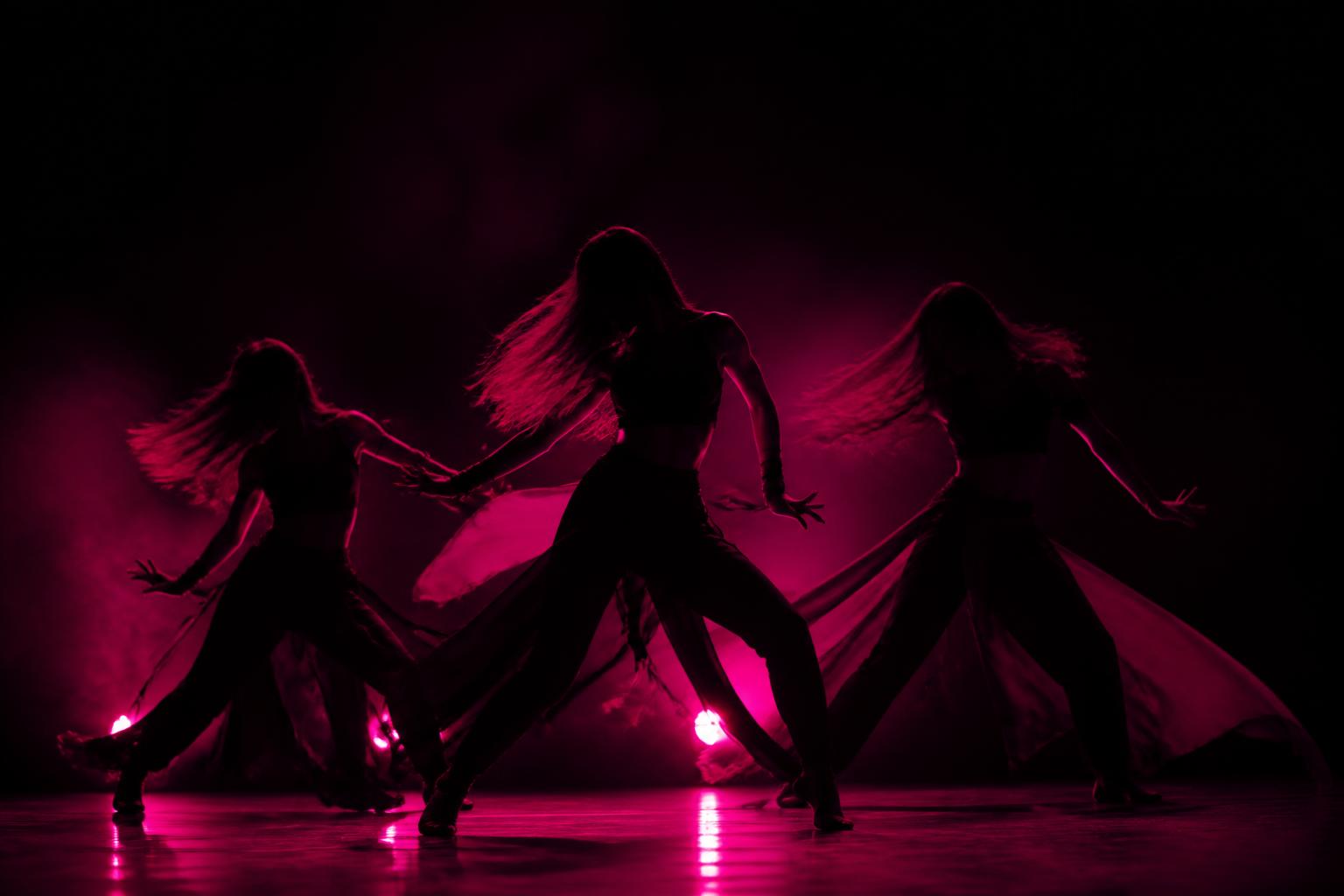 Three dancers in dark outfits under electric pink spotlights